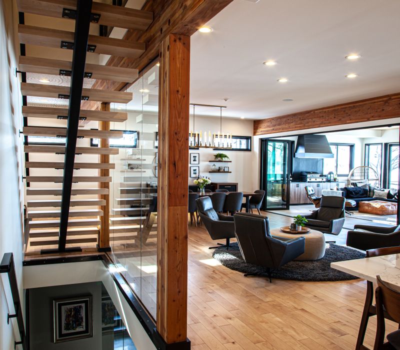 This mid-century modern home includes dramatic central stringer stairs behind a glass wall, white chevron tiles with dark grout, retro furniture, brushed gold fixtures, black cabinets, and laminated beams. 
