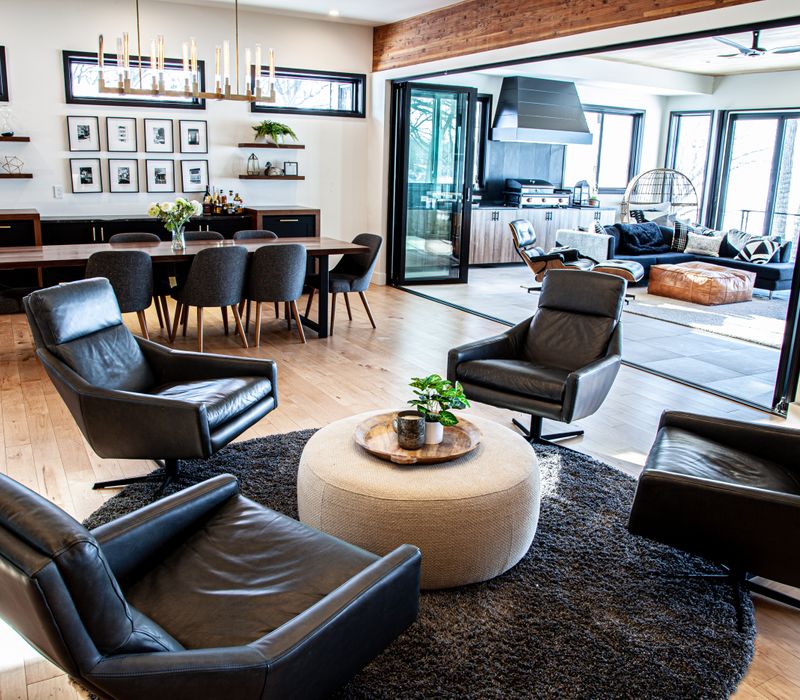 This mid-century modern home includes dramatic central stringer stairs behind a glass wall, white chevron tiles with dark grout, retro furniture, brushed gold fixtures, black cabinets, and laminated beams. 