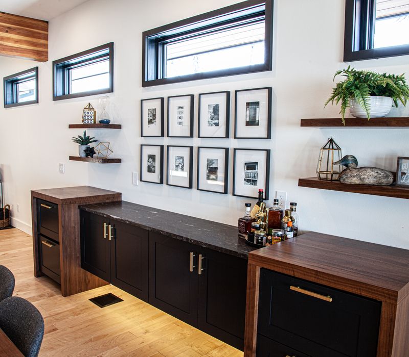 This mid-century modern home includes dramatic central stringer stairs behind a glass wall, white chevron tiles with dark grout, retro furniture, brushed gold fixtures, black cabinets, and laminated beams. 