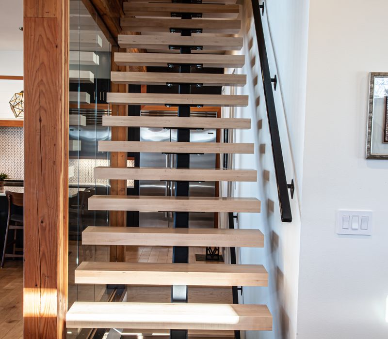 This mid-century modern home includes dramatic central stringer stairs behind a glass wall, white chevron tiles with dark grout, retro furniture, brushed gold fixtures, black cabinets, and laminated beams. 