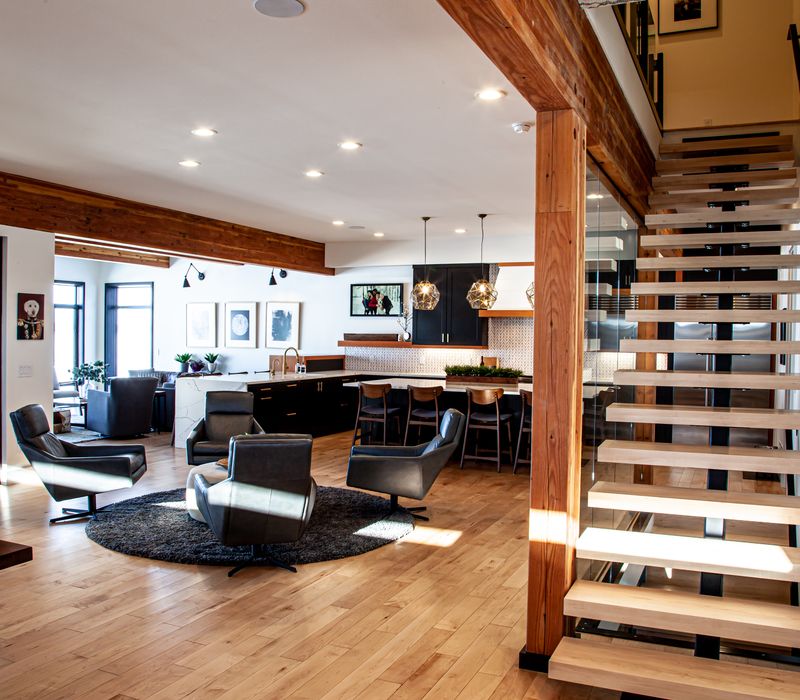 This mid-century modern home includes dramatic central stringer stairs behind a glass wall, white chevron tiles with dark grout, retro furniture, brushed gold fixtures, black cabinets, and laminated beams. 