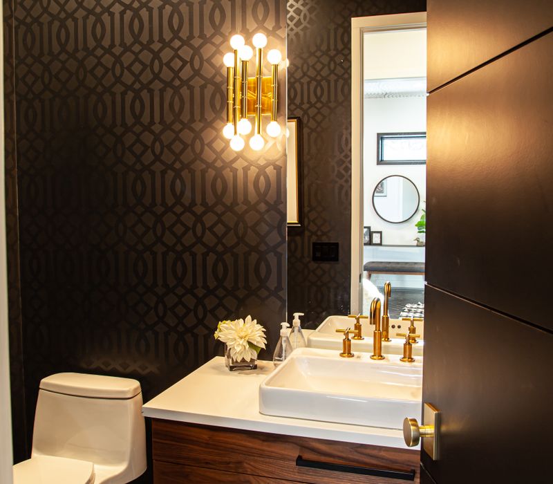 This mid-century modern home includes dramatic central stringer stairs behind a glass wall, white chevron tiles with dark grout, retro furniture, brushed gold fixtures, black cabinets, and laminated beams. 