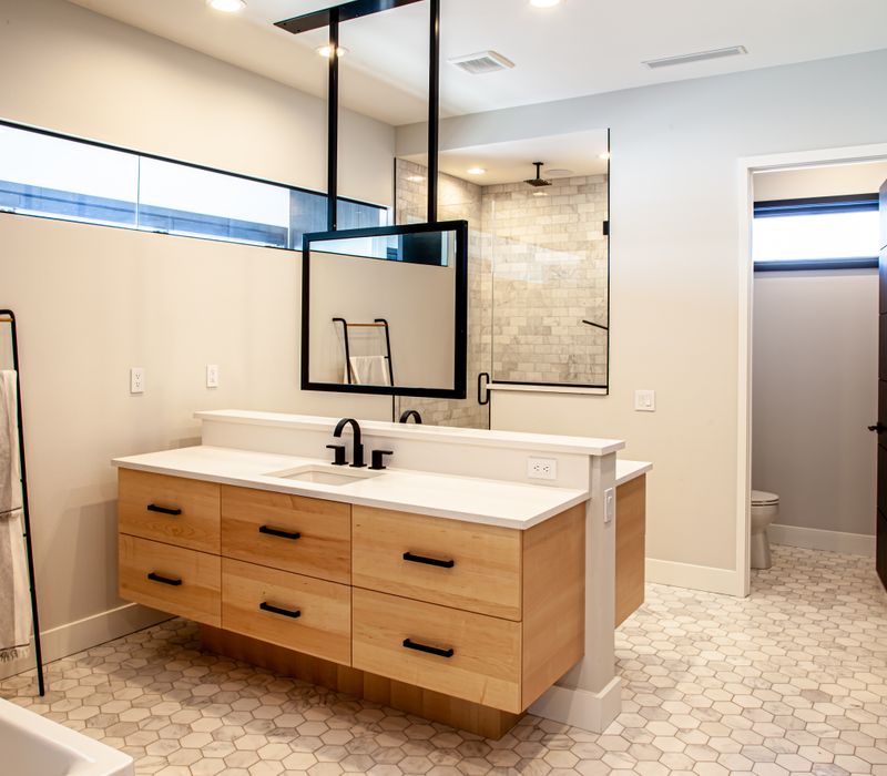 This mid-century modern home includes dramatic central stringer stairs behind a glass wall, white chevron tiles with dark grout, retro furniture, brushed gold fixtures, black cabinets, and laminated beams. 