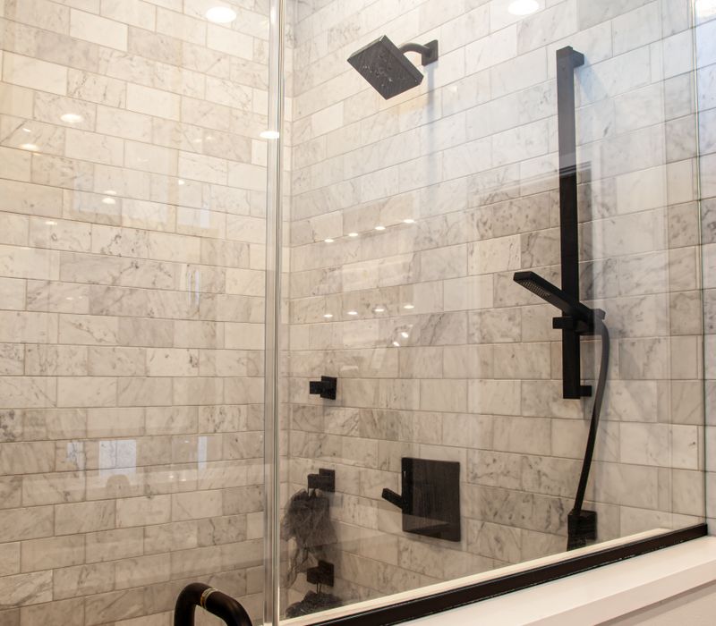 This mid-century modern home includes dramatic central stringer stairs behind a glass wall, white chevron tiles with dark grout, retro furniture, brushed gold fixtures, black cabinets, and laminated beams. 