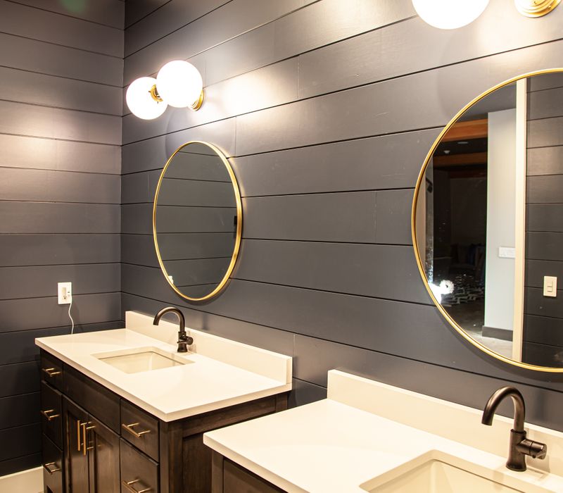 This mid-century modern home includes dramatic central stringer stairs behind a glass wall, white chevron tiles with dark grout, retro furniture, brushed gold fixtures, black cabinets, and laminated beams. 