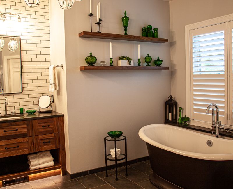 A retro inspired bathroom with a black and white freestanding tub, green glass accents, white subway tiles with dark grout, chrome fixtures, and walnut cabinetry.
