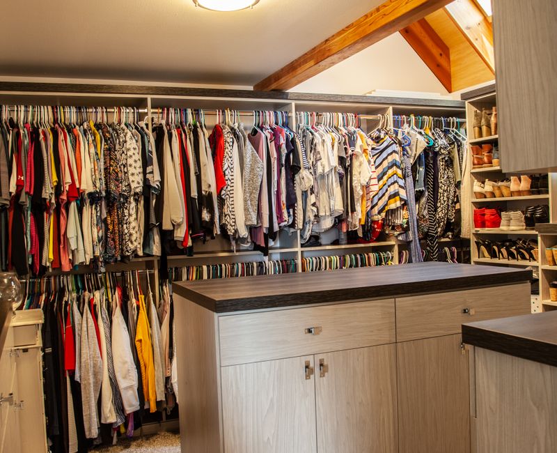 This walk-in closet features wooden ceiling beams that coordinate with the rest of the house, as well as a skylight, central counter, and lots of storage space in the modern cabinets.