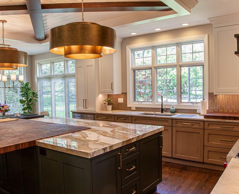 This warm, inviting kitchen features modern brushed gold chandeliers, marble countertops with gold veins, an end-grain cutting board, a metallic glass tile backsplash, and a black range hood with brass accents.
