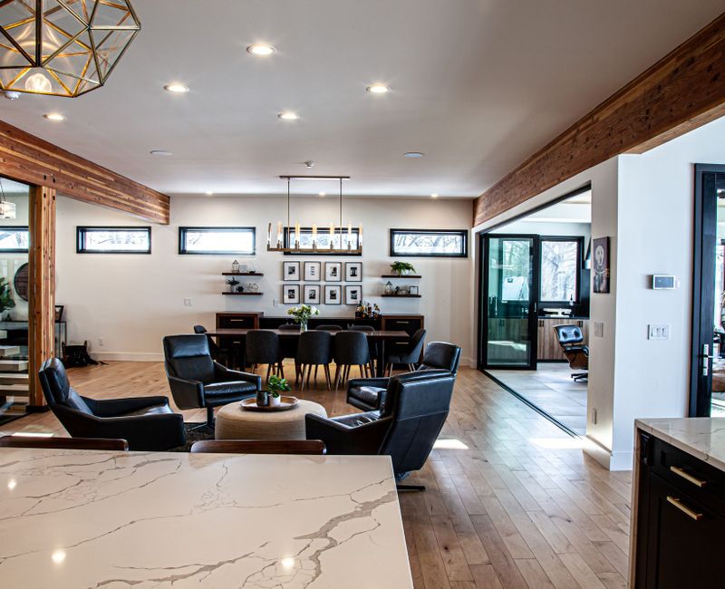 This mid-century modern home includes dramatic central stringer stairs behind a glass wall, white chevron tiles with dark grout, retro furniture, brushed gold fixtures, black cabinets, and laminated beams. 